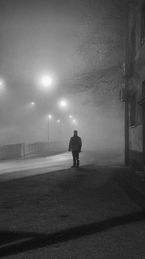 A lone figure standing on a foggy street under street lamps